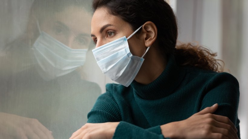 sad young woman wearing face mask looking out of a window