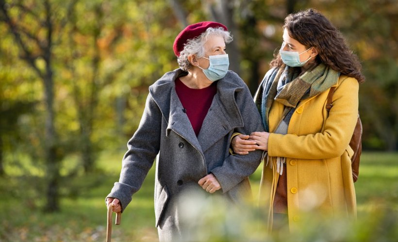 elderly woman walking in park with younger woman. both are wearing a face mask...