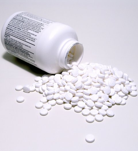 A container for medical pills lies open on a white surface. In front of the round box, many white pills are scattered