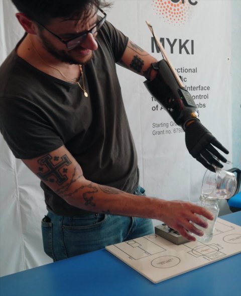 A man with a robotic arm stands at a table. He uses the robotic arm to pour water from one glass into another