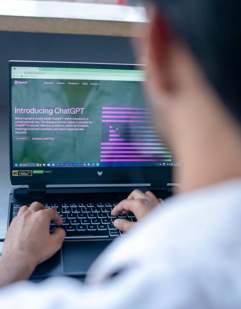 Looking over the shoulder of a man in a white shirt, who is typing on a laptop PC. The screen shows a welcome message from the ChatGPT website