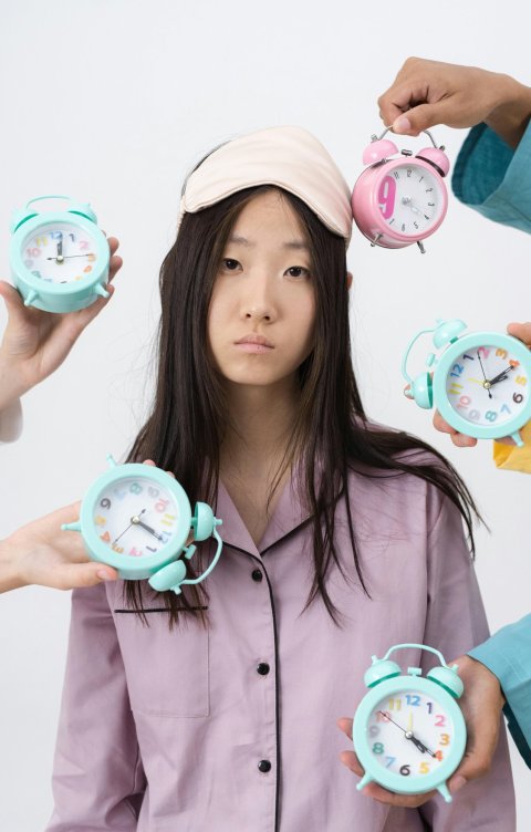 A tired-looking woman wearing a sleeping mask is surrounded by alarm clocks held by hands