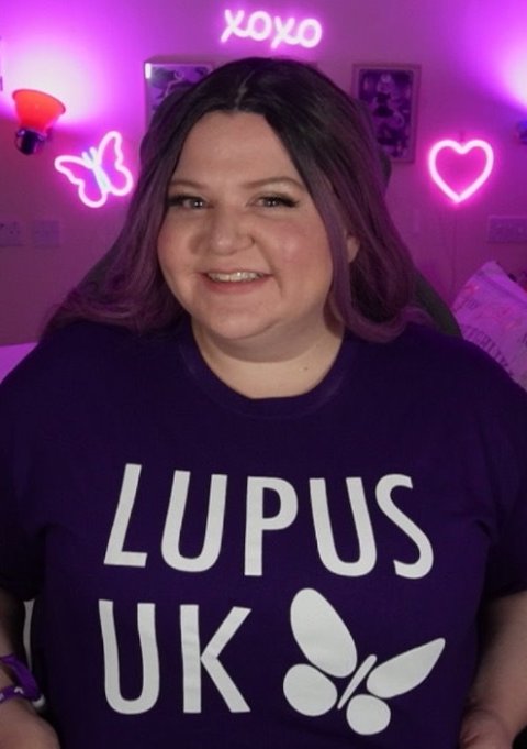 A woman wearing a shirt saysing Lupus UK, sitting in a room with neon light decoration