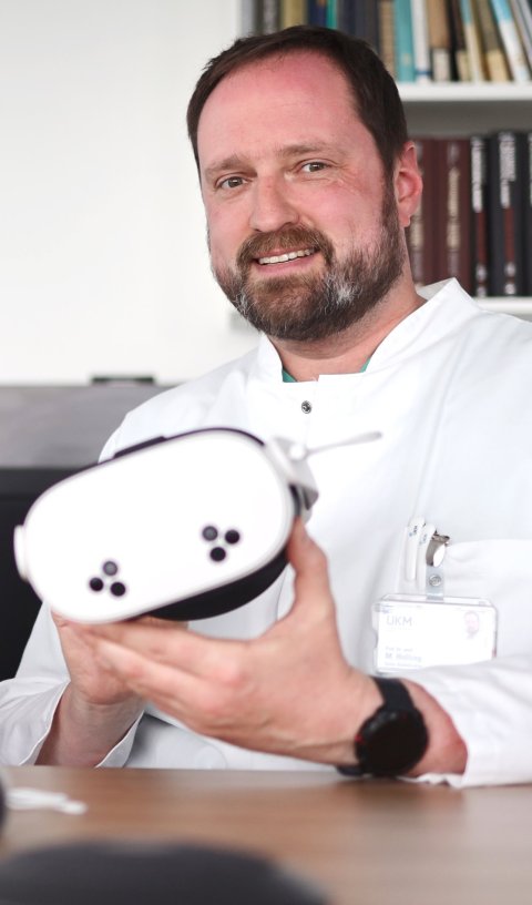 Prof. Markus Holling sitzt an einem Holztisch und hält ein VR-Headset in seinen Händen