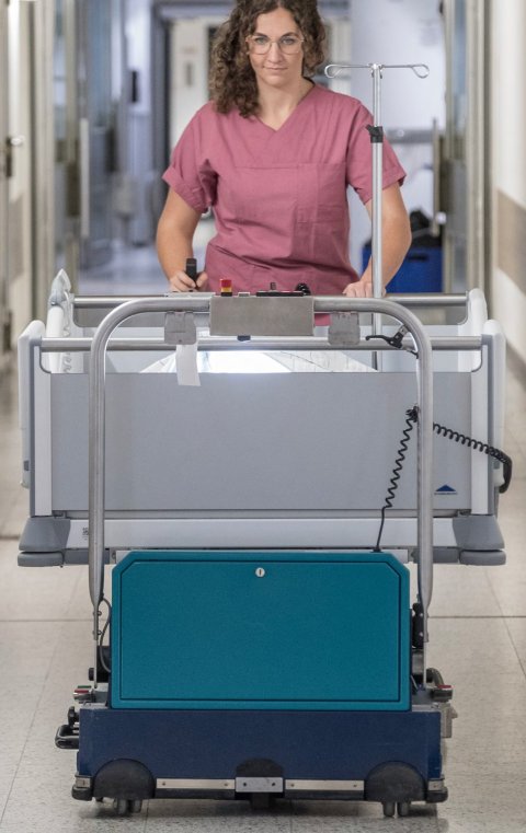 Eine Frau im rosa Kasack schiebt ein Krankenhausbett über einen Krankenhausflur. Ein Gerät zur Unterstützung ist an das Bett gekoppelt