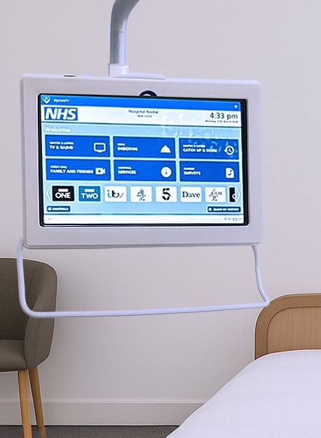 A monitor with a white handle is hanging adjacent to a patient bed. The screen features the NHS logo and a user interface in blue and white