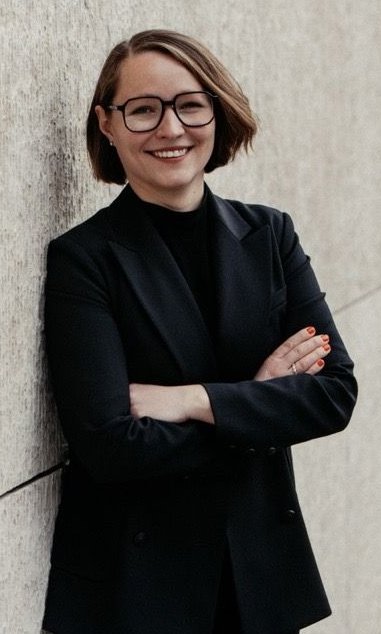Carmen Berger lehnt mit dem Rücken an einer grauen Wand und lächelt mit vor der Brust verschränkten Armen in die Kamera