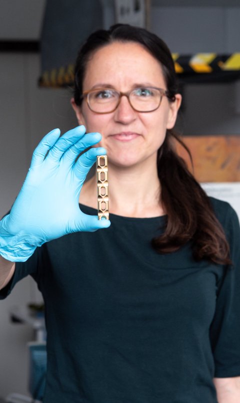 Martina Cihova hält mit der rechten Hand, die in einem blauen Gummihandschuh steckt, ein kleines metallisches Objekt in die Kamera