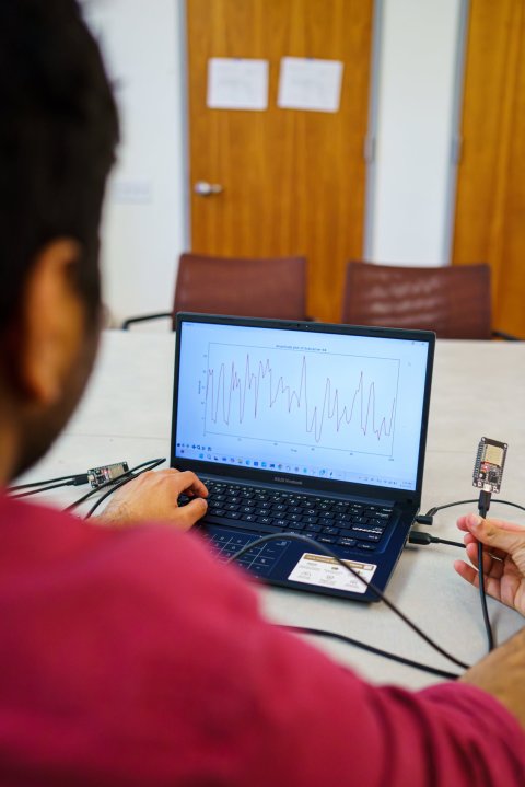 A person in a red sweater is sitting in front of a laptop. On the screen, curves of a heart beat can be seen. The man is holding a small electronic device in his right hand