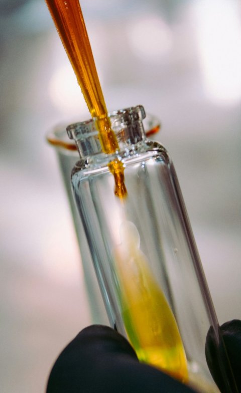 A glass test tube in a laboratory is filled with a yellow liquid from a pipette. the tube is held by a hand in a black rubber glove