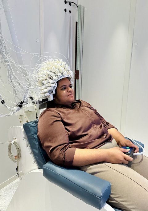 Eine Frau sitzt auf einem Stuhl und trägt einen großen Helm mit vielen Elektroden, die in einem dicken Kabelstrang nach oben links verbunden sind