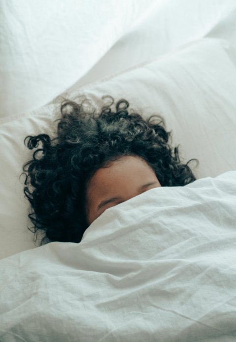 A woman's hair and forehead are peeking out between a bed's blanked and pillow