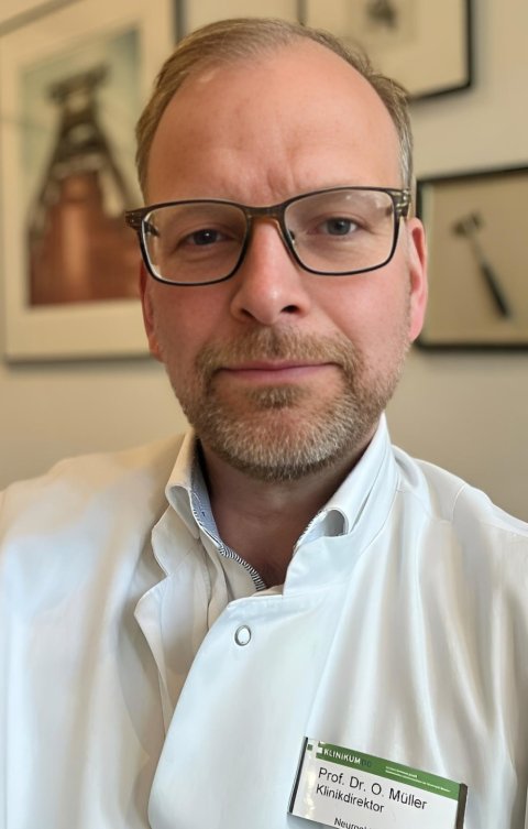 Portraitfoto von Prof. Oliver Müller. Im Hintergrund sind unscharf mehrere gerahmt Bilder zu sehen, unter anderem von der Zeche Zollverein in Essen