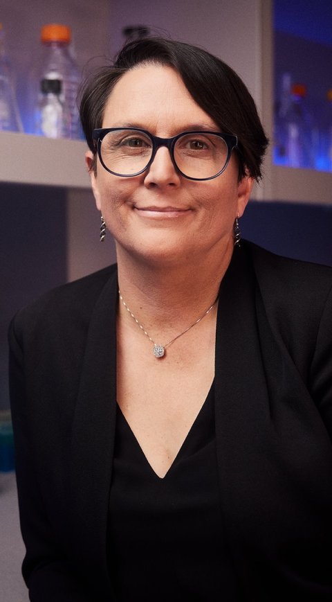 Photo of Aimée Dudley, PhD in a laboratory setting, with different glass flasks in the background
