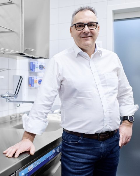 Niklaus Gähler steht in einem Raum für hygieneaufgaben eines Krankenhauses. Seine rechte Hand stützt sich auf eine metallene Arbeitsfläche abgelegt