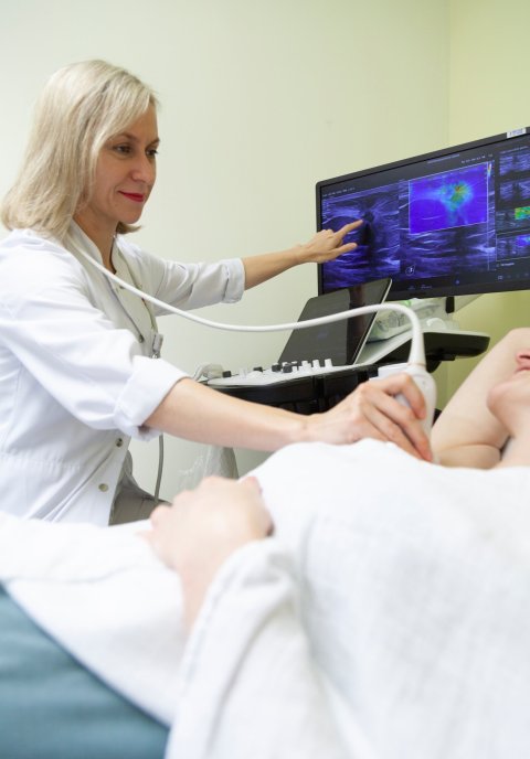 Eine Ärztin untersucht mit per Ultraschall die Lymphknoten einer Patientin. Mit der rechten hand hält sie die Ultraschall-Sonde and die Achsel der Frau, mit der linken zeigt sie auf den Monitor des Ultraschallgeräts