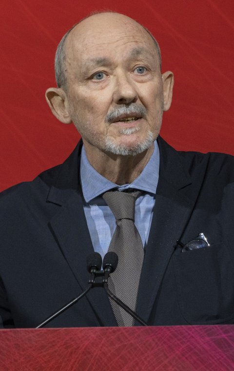 Dr Angel Chamorro is standing at a speaker's podium, addressing his audience. The podium and background are mainly red