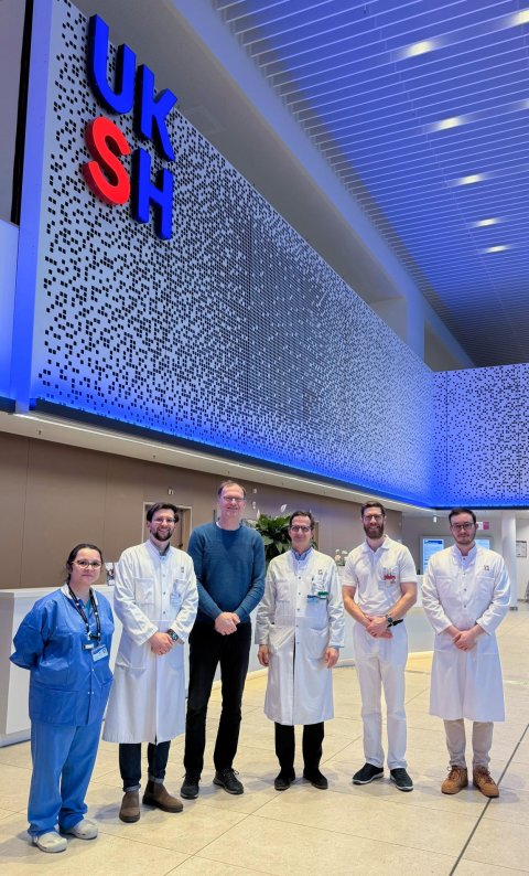 Sechs Personen stehen in der hohen Eingangshalle eines Universitätskrankenhauses mit blauer Deckenbeleuchtung und dem Schriftzug UKSH