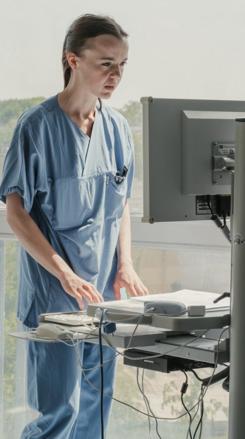 Eine Ärztin im blauen Kasack steht vor einem PC-Pult und blickt auf den Monitor vor ihr. Sie bedient die Tastatur und schaut konzentriert