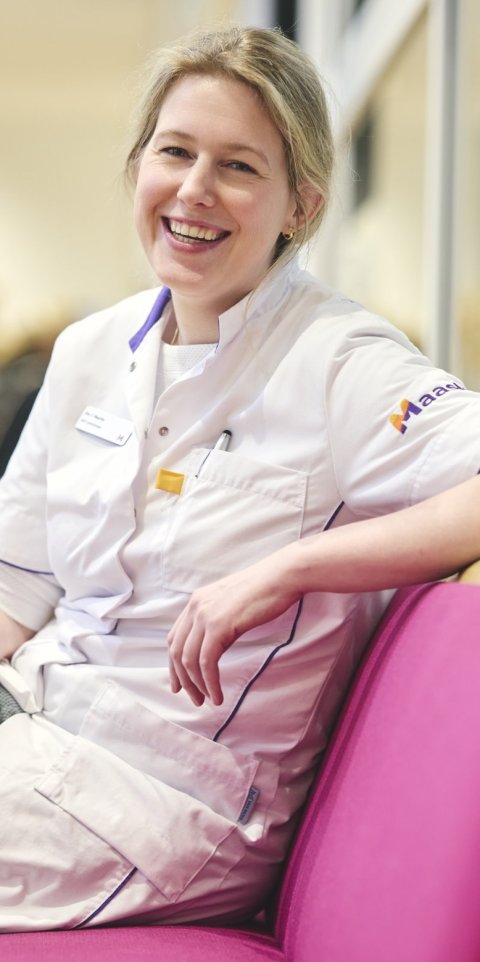 Dr Fleur Mauritz is sitting on a bright pink bench in a hospital. Her left arm is rested on the backrest, and she is smiling at the camera