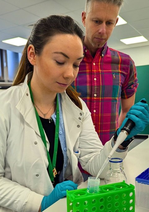 Dr Caitlin O'Shea and Dr Gareth Wright in a laboratory. O'shea is operating a pipette to dispense liquid into a test tube