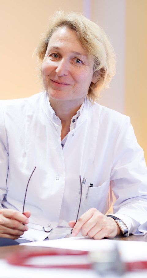 Anke Reinacher-Schick sitzt an einem Screibtisch und hält ihre Brille in beiden Händen