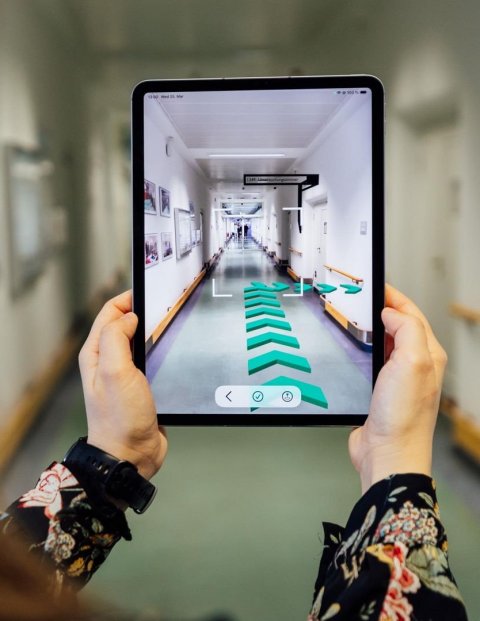 Eine Frau hält ein Tablet hoch, das in einer Augmented-Reality-Ansicht Pfeile zur Navigation in einem Krankenhausflur anzeigt