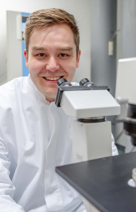 Johannes Karges sitzt im weißen Laborkittel hinter einem Mikroskop mit Objektträger