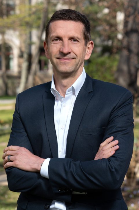 André Rauschert steht mit vor der Brust verschränkten Armen in einem sonnigen Park