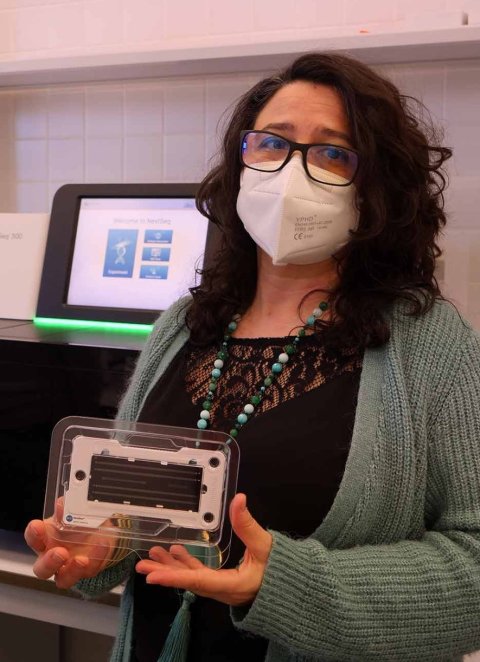 Prof. Ana Grande is standing in a medical laboratory environment wearing a protective face mask. She is holding a small device in both hands