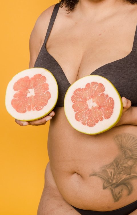 Cropped photo of an overweight woman against an orange background. The woman is holding the two halves of a grapefruit in front of her breasts