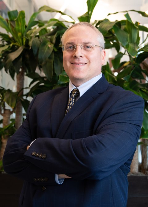 Dr Liron Pantanowitz is standing in a suit with his arms crossed in front of his chest. In the background, a huge indoor plant can be seen