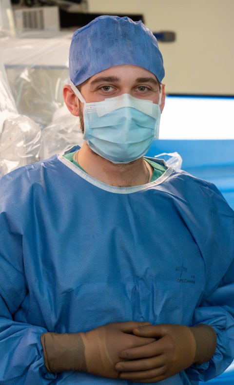 Dr. Julius Kaemmel in blauer OP-Kleidung mit Haube und Mundschutz vor einem Operationsroboter