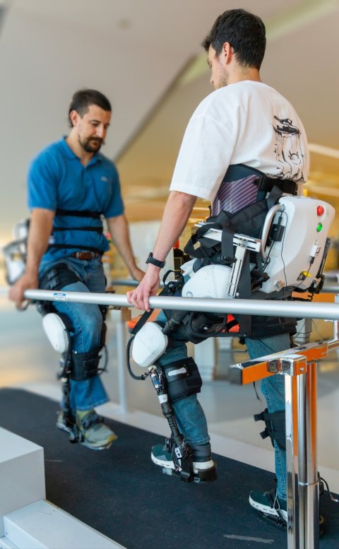 Two men are wearing lower-limb exoskeletons on their legs for gait training on a pair of handlebars