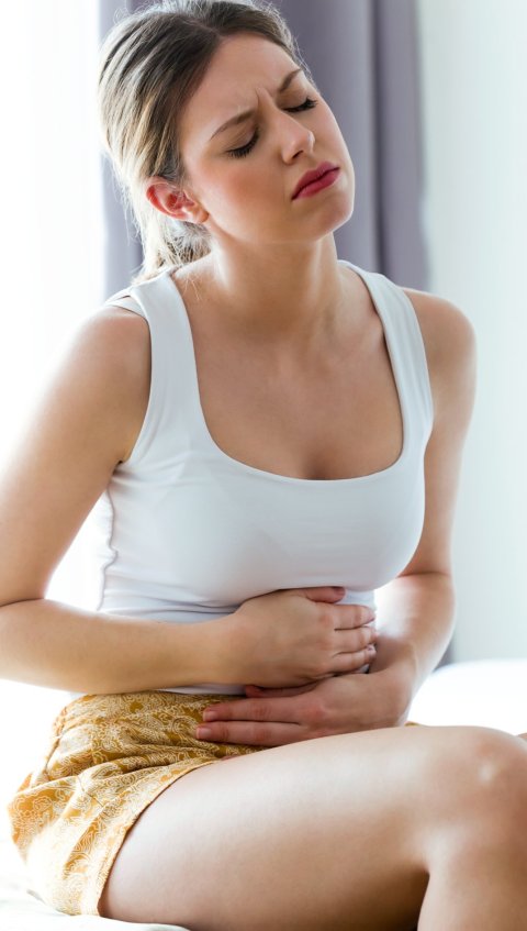A young woman is sitting on a sofa, holding her stomach from pain