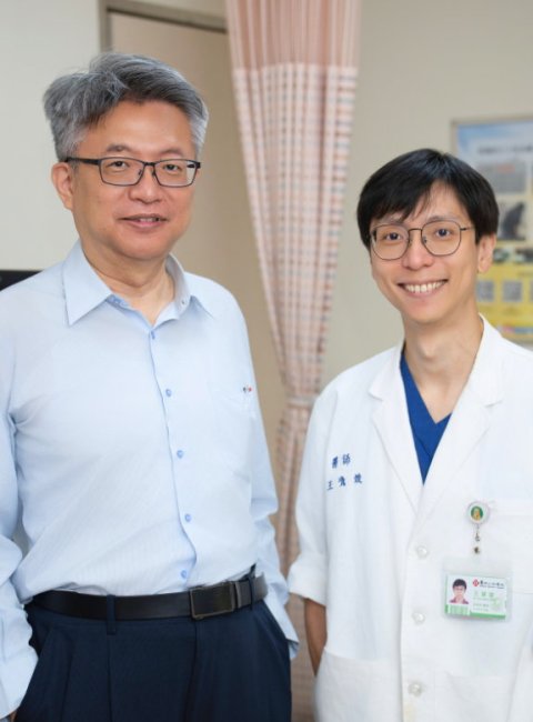 Two men are standing next to each other in a medical examination room, looking at the camera and smiling