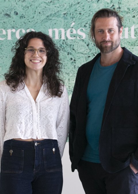 Dr. Alejandra Morcillo-Nieto and Dr. Alexandre Bejanin are standing in front of a wall with a green picture of cancerous tissue and the text Més recerca, més salut (More research, better health), the motto of IR Sant Pau