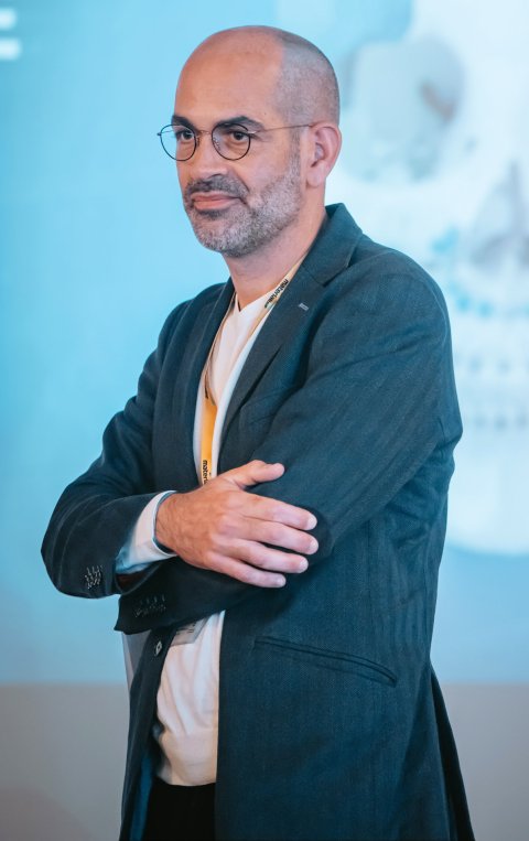 Prof. Thomas Schouman is standing with his arms crosed in front of his chest. In the background, an image of a giant human skull can be seen
