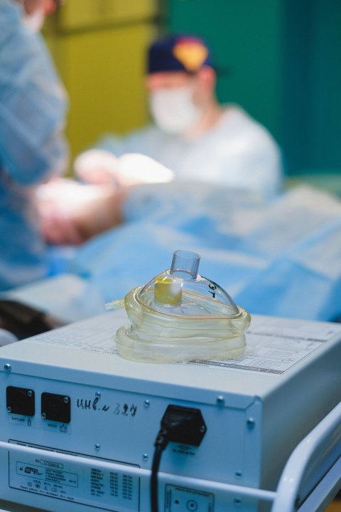 Two transparent mouthpieces used for anaesthesia are lying on top of a gray device in a hospital's operating theatre. In the background, a surgeon is performing surgery